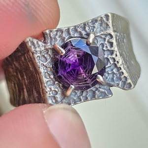 May include: A silver ring with a textured band and a large, faceted, deep purple gemstone. The gemstone is held in place by four silver prongs. The ring has a unique, artistic design.