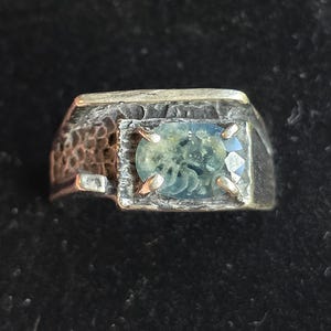 May include: A silver ring with a rectangular setting. The setting holds a light blue gemstone with a faceted cut. The band has a textured, hammered appearance, adding a rustic touch to the jewelry.
