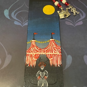 May include: A black wooden bookmark with a painted scene of a clown standing in front of a red and white striped circus tent under a full moon. The bookmark has a red ribbon and silver charm.