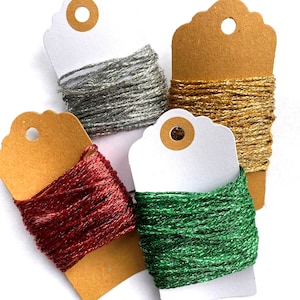 May include: Four spools of metallic thread on brown and white tags. The thread colours are silver, gold, red, and green.