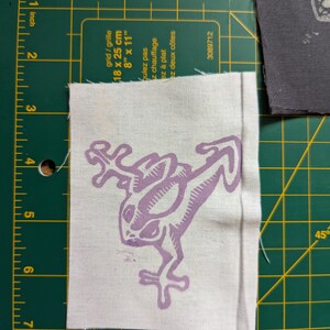 May include: A white fabric patch with a light purple frog design. The frog is stylized with long limbs and a detailed body. The patch is approximately 3 inches by 3 inches. The patch is on a green cutting mat with inch and centimeter measurements.
