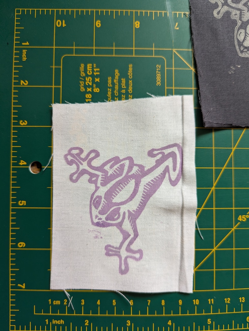 May include: A white fabric patch with a light purple frog design. The frog is stylized with long limbs and a detailed body. The patch is approximately 3 inches by 3 inches. The patch is on a green cutting mat with inch and centimeter measurements.