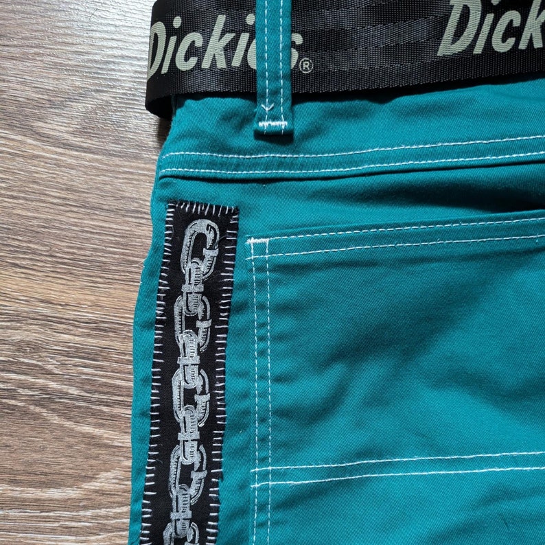 Peut inclure: Pantalon de couleur teal avec des coutures blanches et une ceinture noire avec le logo Dickies. Un patch noir avec un motif de cha&icirc;ne est cousu sur le c&ocirc;t&eacute;. Le pantalon est en tissu r&eacute;sistant.