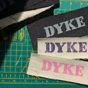 Könnte beinhalten: Mehrere Stoffetiketten mit dem Wort "DYKE" in verschiedenen Farben, darunter Schwarz, Lila und Rosa. Die Etiketten sind rechteckig und scheinen handgefertigt zu sein, mit sichtbaren ausgefransten Kanten. Der Hintergrund ist eine grüne Schneidematte.