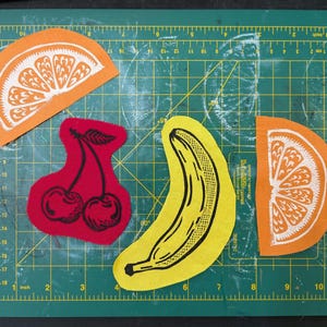 Op de afbeelding: Een verzameling stoffen fruituitsparingen, waaronder twee sinaasappelschijfjes, een banaan en een paar kersen. De sinaasappelschijfjes zijn oranje met witte details, de banaan is geel met zwarte details en de kersen zijn rood met zwarte details.
