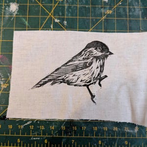 Può includere: Stampa linoleografica in bianco e nero di un uccello su un pezzo di tessuto bianco. L'uccello è appollaiato su un ramo, con dettagliati motivi di piume. Il tessuto è rettangolare con bordi grezzi, su un tappetino da taglio verde.
