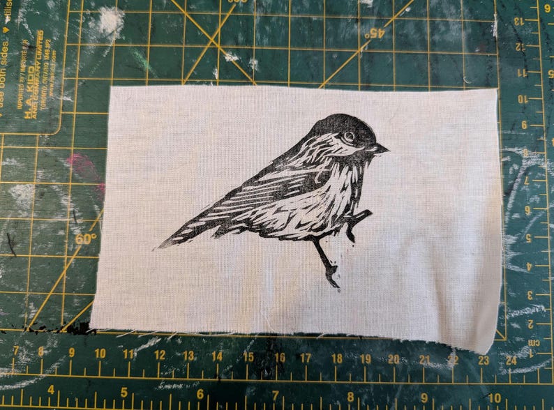 Op de afbeelding: Een zwart-wit linoleumsnede van een vogel op een stuk witte stof. De vogel zit op een tak, met gedetailleerde veermotieven. De stof is rechthoekig met rafelige randen, tegen een groene snijmat.