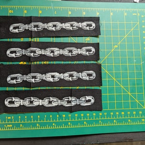 May include: Four black fabric patches featuring a silver chain design. The patches are rectangular and arranged on a green cutting mat. The chain links are intricately embroidered, creating a striking contrast against the black fabric.