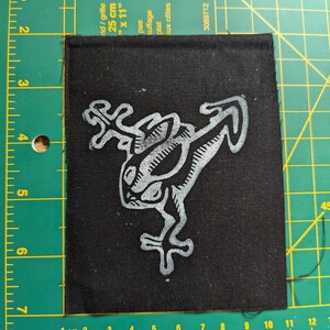 May include: Black square patch with a white stylized frog design. The patch is approximately 3 inches by 3 inches. The frog illustration has a unique, artistic style, with detailed lines and shapes. The patch is likely for clothing or accessories.
