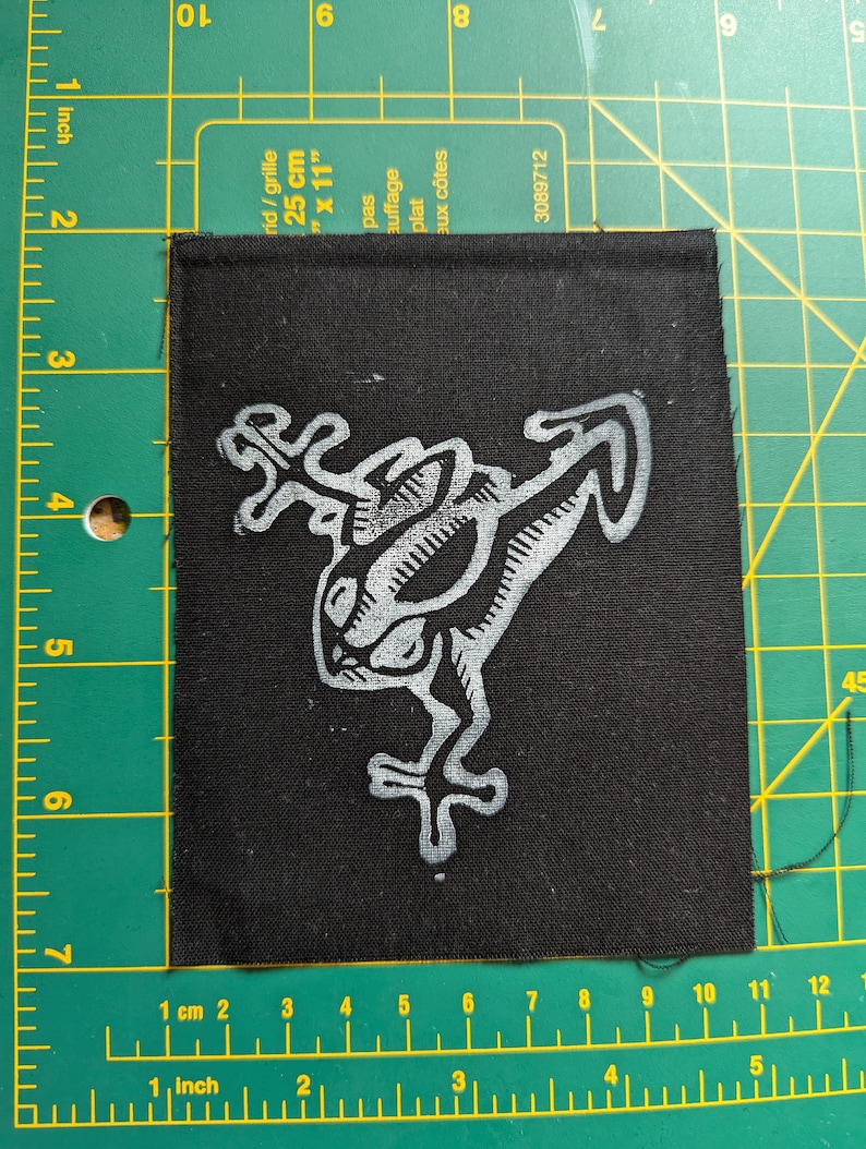 May include: Black square patch with a white stylized frog design. The patch is approximately 3 inches by 3 inches. The frog illustration has a unique, artistic style, with detailed lines and shapes. The patch is likely for clothing or accessories.
