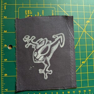 May include: A dark gray fabric patch featuring a stylized, light gray frog design. The frog has large eyes and outstretched limbs. The patch is rectangular with a stitched border and is approximately 3 inches by 3 inches.