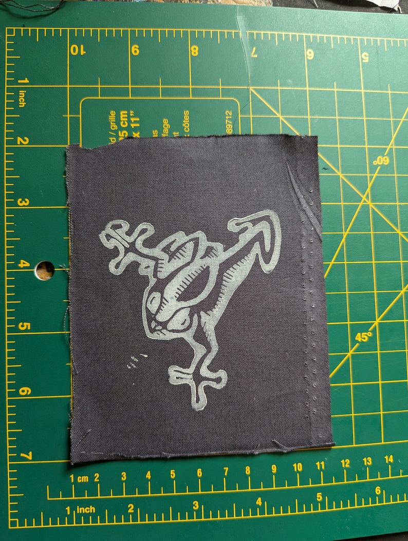 May include: A dark gray fabric patch featuring a stylized, light gray frog design. The frog has large eyes and outstretched limbs. The patch is rectangular with a stitched border and is approximately 3 inches by 3 inches.