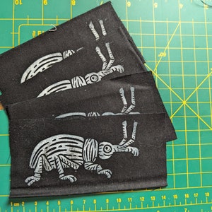 May include: A stack of black fabric squares, each featuring a light grey insect print. The insect design includes a segmented body, legs, and antennae. The squares are on a green cutting mat with cm measurements.