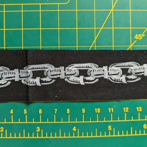 Peut inclure: Un &eacute;cusson en tissu noir avec un motif de cha&icirc;ne argent&eacute;e brod&eacute;e. Le patch rectangulaire mesure environ 15 cm de long et 4 cm de large. L'&eacute;cusson est pos&eacute; sur un tapis de coupe vert.