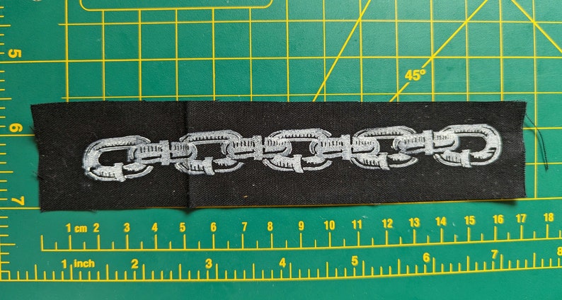 Peut inclure: Un &eacute;cusson en tissu noir avec un motif de cha&icirc;ne argent&eacute;e brod&eacute;e. Le patch rectangulaire mesure environ 15 cm de long et 4 cm de large. L'&eacute;cusson est pos&eacute; sur un tapis de coupe vert.