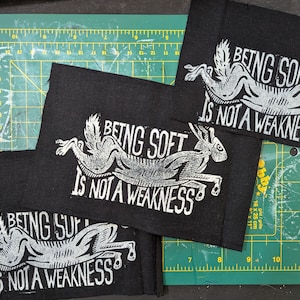 May include: Black fabric patches featuring a white illustration of a running hare and the text "BEING SOFT IS NOT A WEAKNESS". The patches are rectangular and rest on a green cutting mat, approximately 18 x 25 cm.