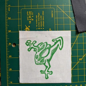 May include: A square, off-white fabric patch featuring a green frog design. The frog is stylized with a linocut aesthetic. The patch is approximately 4 inches by 4 inches. The patch is on a green cutting mat with grid lines.
