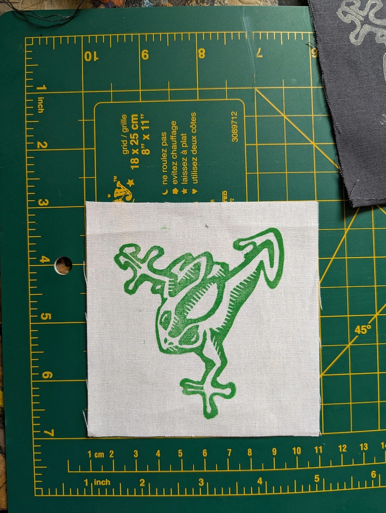May include: A square, off-white fabric patch featuring a green frog design. The frog is stylized with a linocut aesthetic. The patch is approximately 4 inches by 4 inches. The patch is on a green cutting mat with grid lines.