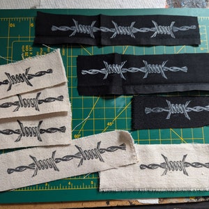 May include: Various fabric patches featuring a barbed wire design and the word "Jooor" printed in a stylized font. The patches are in different sizes and colors, including black and beige. The patches are laid out on a green cutting mat.