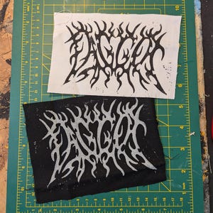 May include: Two fabric patches with the word "DAGGER" in a tribal-style font. One patch is white with black text, and the other is black with silver text. The patches are rectangular and sit on a green cutting mat.