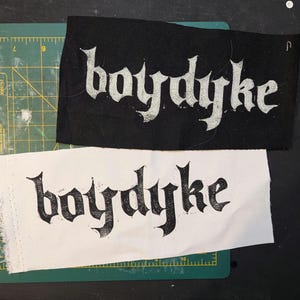 May include: Two fabric patches, one black and one white, each bearing the word "boydyke" in a gothic font. The black patch has white text, the white patch has black text. They rest on a green cutting mat and a black surface.