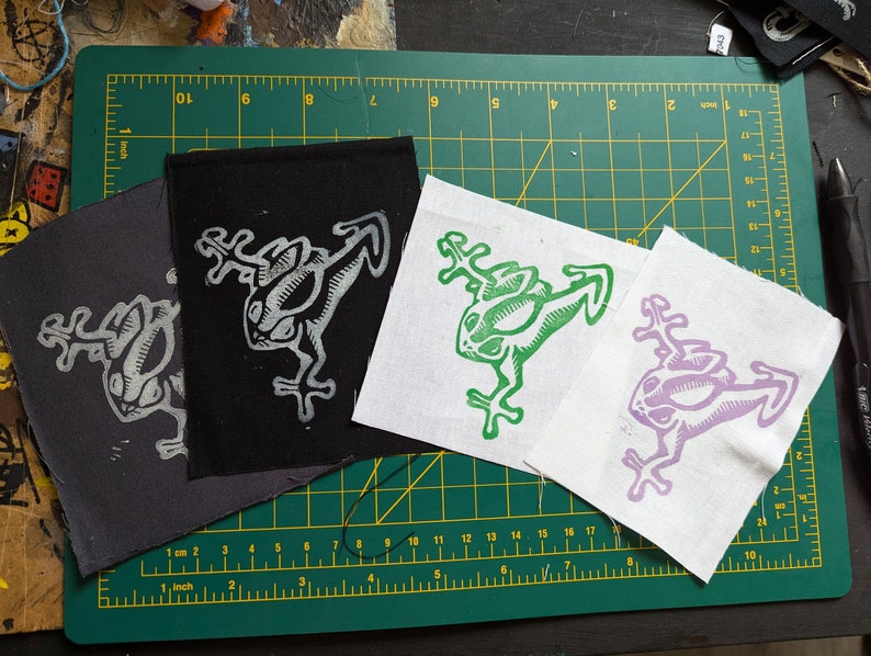 May include: Four fabric patches with a frog design. The patches are in shades of gray, black, green, and purple. The frog design is a stylized illustration. The patches are on a green cutting mat.