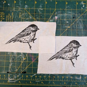 Op de afbeelding: Twee stoffen vierkanten met een zwart-wit vogelprint. De vogel is afgebeeld in een gedetailleerde linosnede stijl, met de nadruk op zijn veren en houding. De stof heeft een natuurlijke, gebroken witte kleur.