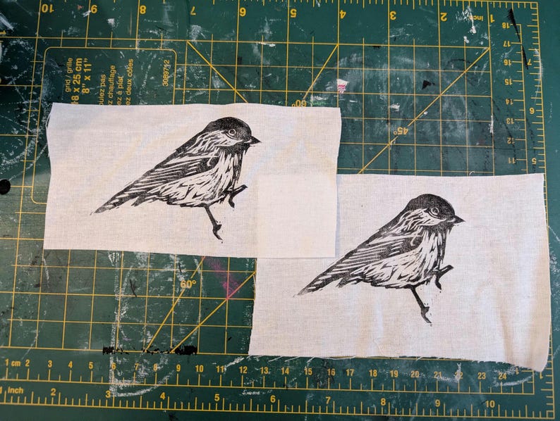 Op de afbeelding: Twee stoffen vierkanten met een zwart-wit vogelprint. De vogel is afgebeeld in een gedetailleerde linosnede stijl, met de nadruk op zijn veren en houding. De stof heeft een natuurlijke, gebroken witte kleur.