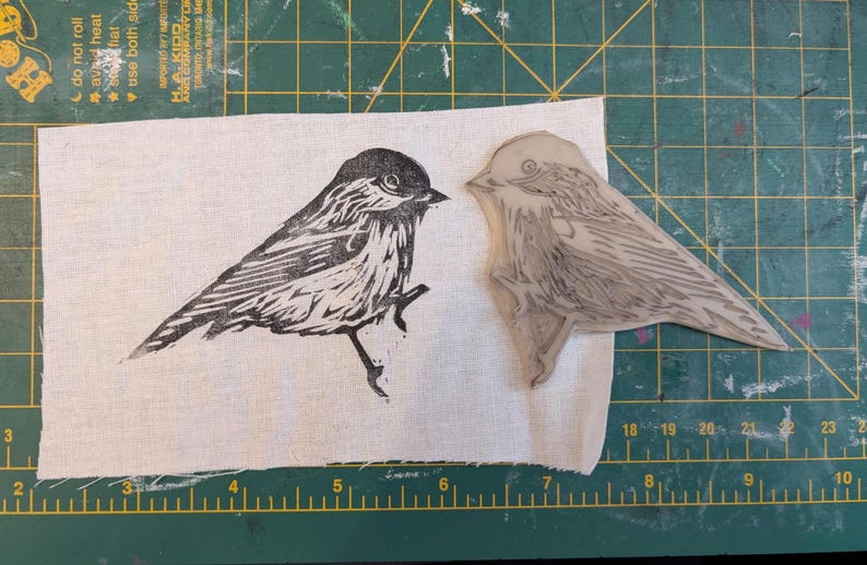 Op de afbeelding: Een zwart-wit linoleumsnede van een vogel op stof, naast het linoleumblok dat werd gebruikt om de afdruk te maken. De vogel zit op een tak. De achtergrond is een groene snijmat.