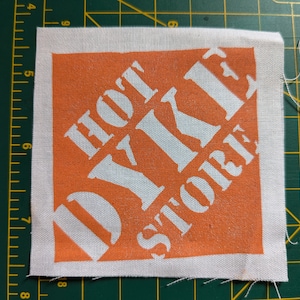 May include: Square fabric patch with an orange background and white text that reads "HOT DIY STORE". The patch has a white border and is placed on a green cutting mat.