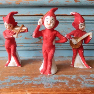 May include: Three ceramic elf figurines painted in bright red with white accents. The left elf plays a violin, the center elf holds a conductor's baton, and the right elf plays a small stringed instrument. Each figure stands on a white base.