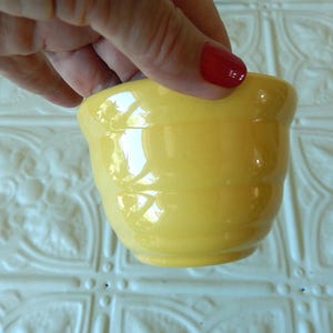 May include: A small, round, yellow ceramic bowl with a glossy finish. The bowl has a slightly flared rim and horizontal ridges around the body. The bowl is held by a hand with red nail polish.
