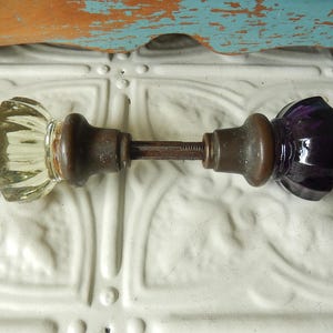 May include: A door knob set featuring two faceted glass knobs, one clear and one deep purple, connected by a central metal spindle. Each knob is attached to a dark brown metal base, showcasing a classic design with multi-sided glass surfaces.