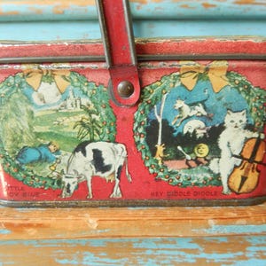 May include: Vintage red metal picnic basket with a red handle. The sides feature colorful illustrations of a cow, a person, a cat playing a cello, and other figures. The text "Little Boy Blue" and "Hey Diddle Diddle" are visible.