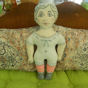 May include: A handmade cloth doll with a painted face, wearing a white romper with blue accents, pink stockings, and gray boots. The doll has light brown hair and is positioned in front of a patterned pillow.