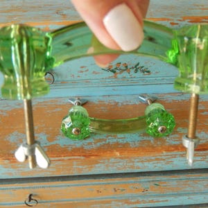 May include: Two translucent lime green glass drawer pulls featuring a curved handle design with faceted ends. Each pull includes two threaded metal bolts extending from the base, secured with silver-toned wing nuts at the bottom of the hardware.