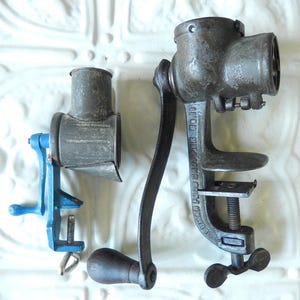 May include: Two antique food choppers. One is a small, blue and silver hand-cranked model. The other is a larger, gray metal chopper with a hand crank and a clamp for securing it to a surface. The larger chopper has the text "ENTERPRISE FOOD CHOPPER NO.10" on the side.