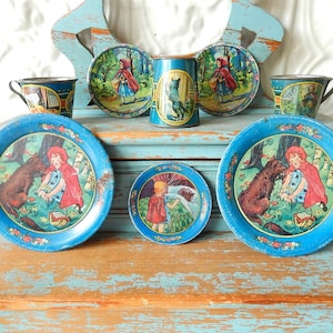 May include: A vintage children's tea set featuring illustrations of Little Red Riding Hood. The set includes blue trays, cups, and a cylindrical container. The artwork depicts the story's characters in a forest setting, with floral borders.