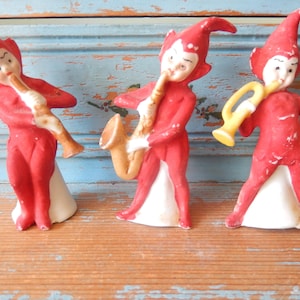 May include: Three ceramic elf figurines dressed in red suits with pointed hats, each playing a different musical instrument. One elf plays a tan flute, another plays a tan saxophone, and the third plays a yellow trumpet, all standing on white bases.