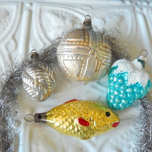 May include: A collection of four vintage glass ornaments including a silver sphere with geometric patterns, a small silver ribbed ornament, a turquoise fruit-shaped ornament with white glitter, and a yellow fish ornament with red accents.