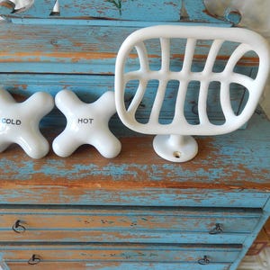 May include: White ceramic bathroom fixtures, including two cross-shaped faucet handles labeled "COLD" and "HOT", and a soap dish. The items are displayed on a distressed blue and brown wooden cabinet, creating a vintage aesthetic.