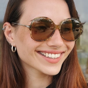 Puede incluir: Una mujer con el pelo largo y castaño que lleva gafas de sol redondas de carey marrón con un aro de nariz plateado.