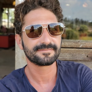 May include: A man with a beard and brown hair is wearing silver-rimmed sunglasses with brown lenses. He is wearing a dark blue shirt.