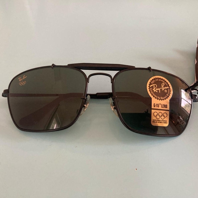 Op de afbeelding: Zwarte Ray-Ban zonnebril met een gouden Olympisch logo op de linkerlens en een gouden sticker op de rechterlens met de tekst "Ray-Ban 100% UV protection G-15 Lens Worldwide Olympic Sponsor"