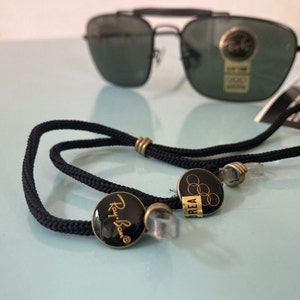 Op de afbeelding: Zwarte brilkoord met gouden Ray-Ban en Olympische ringen bedels.