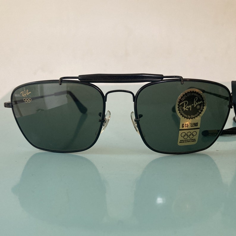 Op de afbeelding: Zwarte Ray-Ban zonnebril met groene glazen en een gouden Olympisch logo op het linker glas. De zonnebril heeft een zwart metalen frame en een zwarte metalen brug. De zonnebril is 100% UV-beschermd en heeft G-15 glazen.