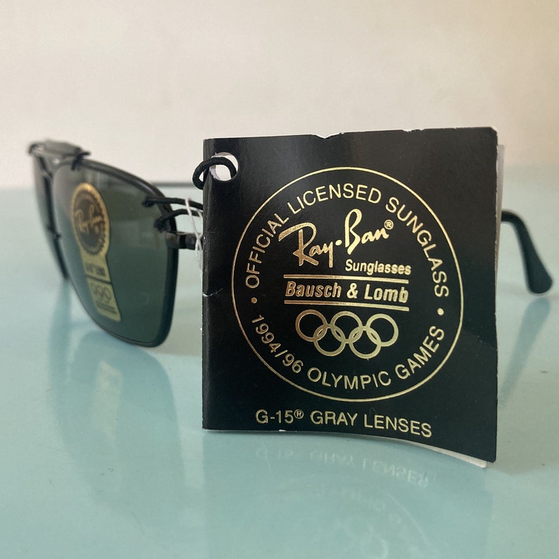 Op de afbeelding: Zwarte Ray-Ban zonnebril met een gouden label met de tekst "OFFICIAL LICENSED SUNGLASSES Ray-Ban Sunglasses Bausch & Lomb 1994/96 OLYMPIC GAMES G-15&reg; GRAY LENSES".