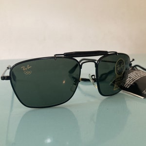 Op de afbeelding: Zwarte Ray-Ban zonnebril met een gouden Olympisch logo op de linkerlens. De zonnebril heeft een opklapbare functie en is in goede staat.