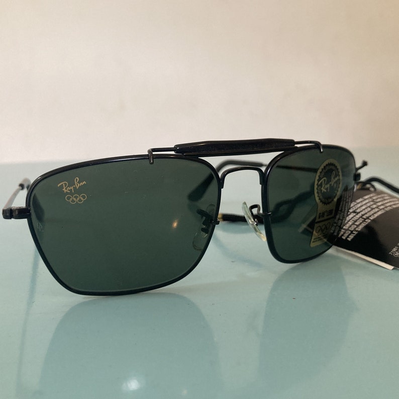 Op de afbeelding: Zwarte Ray-Ban zonnebril met een gouden Olympisch logo op de linkerlens. De zonnebril heeft een opklapbare functie en is in goede staat.
