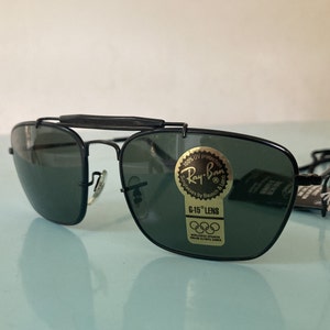 Op de afbeelding: Zwarte Ray-Ban zonnebril met groene glazen en een gouden label met de tekst "100% UV protection Ray-Ban Sunglasses by Bausch & Lomb G-15 Lens Worldwide Sponsor of the Olympic Games".
